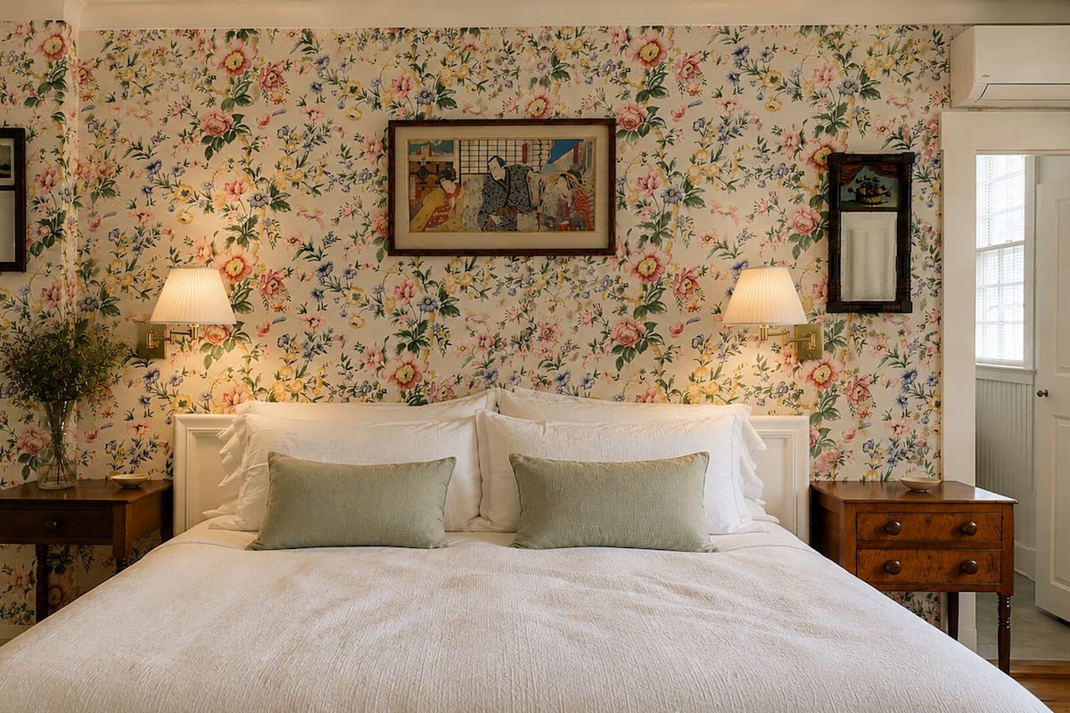 A bedroom with floral wallpaper, a bed with white linens, two bedside tables with lamps, and a framed picture on the wall.