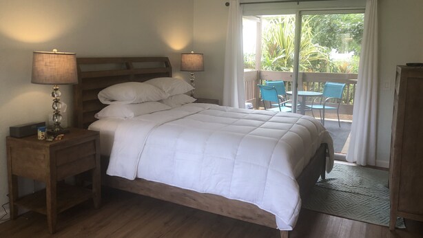 A bedroom with a wooden bed, two bedside lamps, a nightstand, and a view of an outdoor balcony with chairs and plants. - Sandpiper 133B - updated and renovated studio in Princeville  (Princeville)