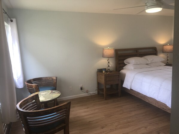 A bedroom with a wooden bed, bedside lamps, a wooden nightstand, a round table with chairs, and a ceiling fan.