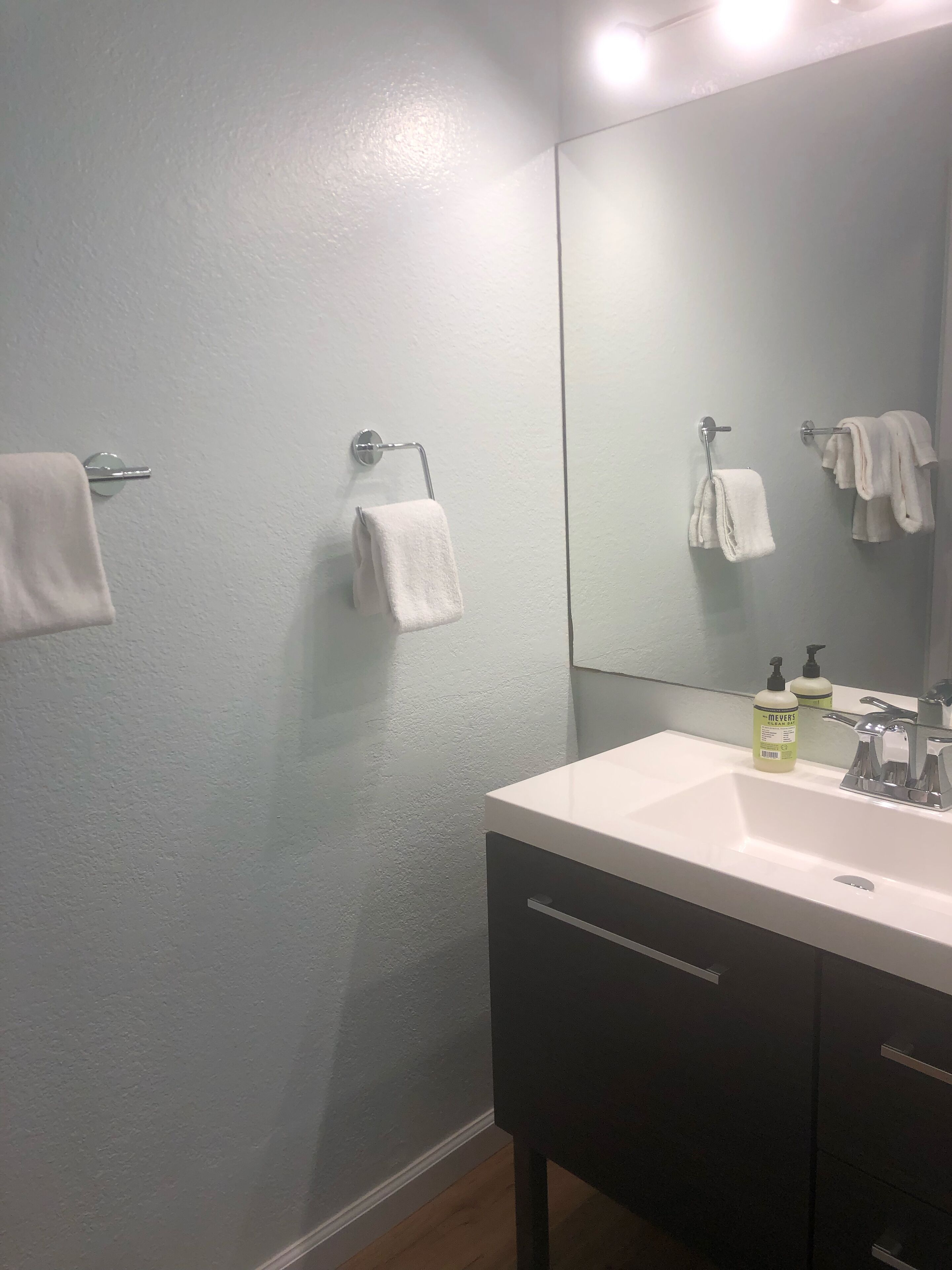 Ein Badezimmer mit einem weißen Waschbecken, dunklen Schränken und einem Spiegel. Zwei Handtücher hängen an Handtuchhaltern und auf dem Waschbecken steht eine Seife.