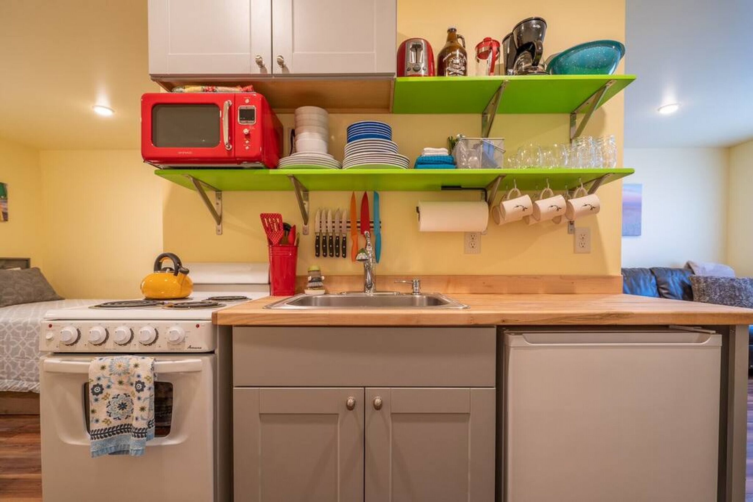 A compact kitchen with a stove, microwave, and sink. There are green shelves above the counter holding dishes and glasses.