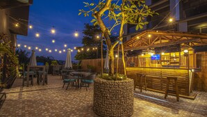 Een terras buiten met betegelde vloer, houten meubels, lichtslingers en een bar met een televisiescherm.