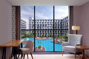 Habitación de hotel con un ventanal grande, vista a la piscina, una silla, una mesa y una lámpara.