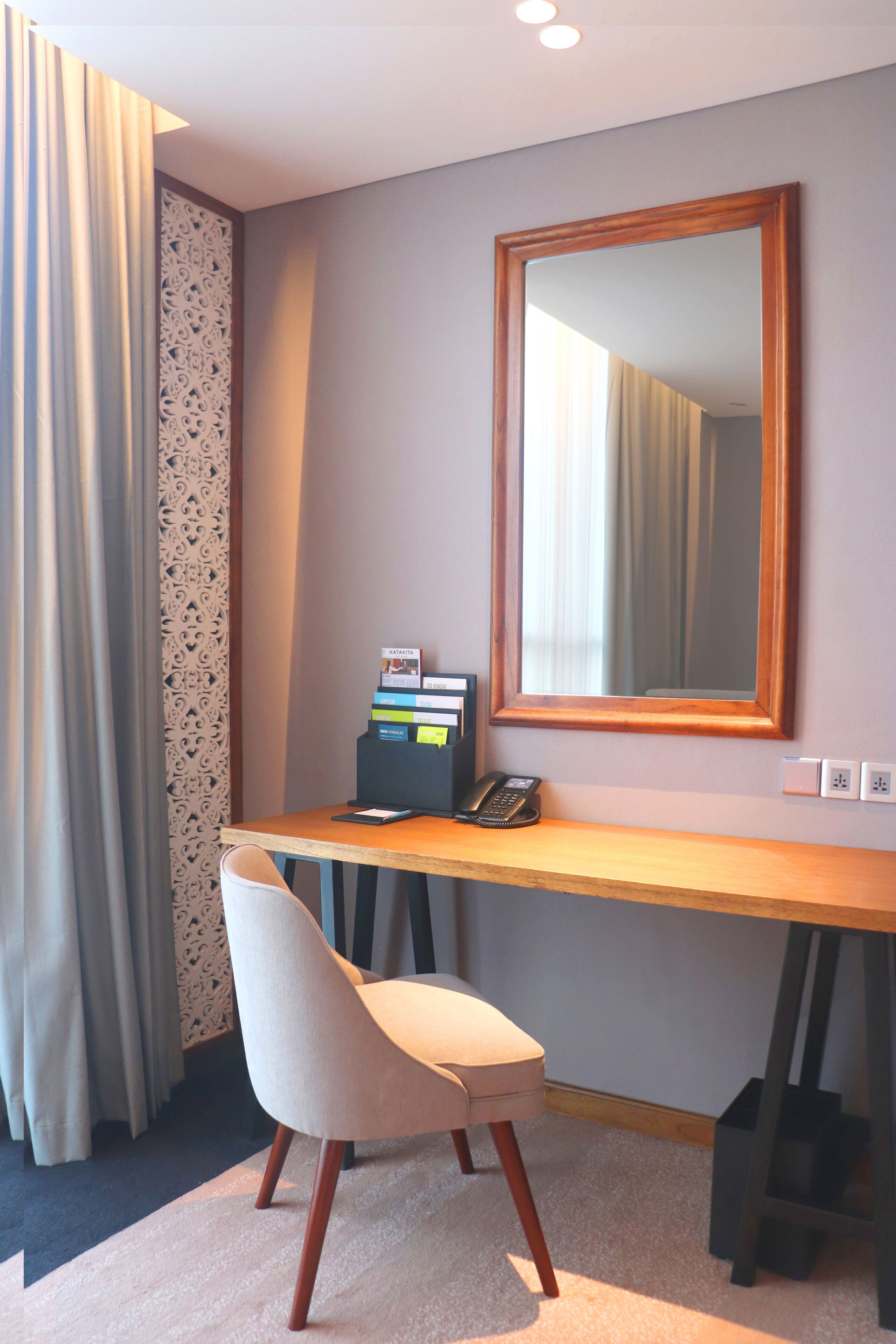 A hotel room with a wooden desk, a chair, a mirror, a telephone, and a decorative panel.