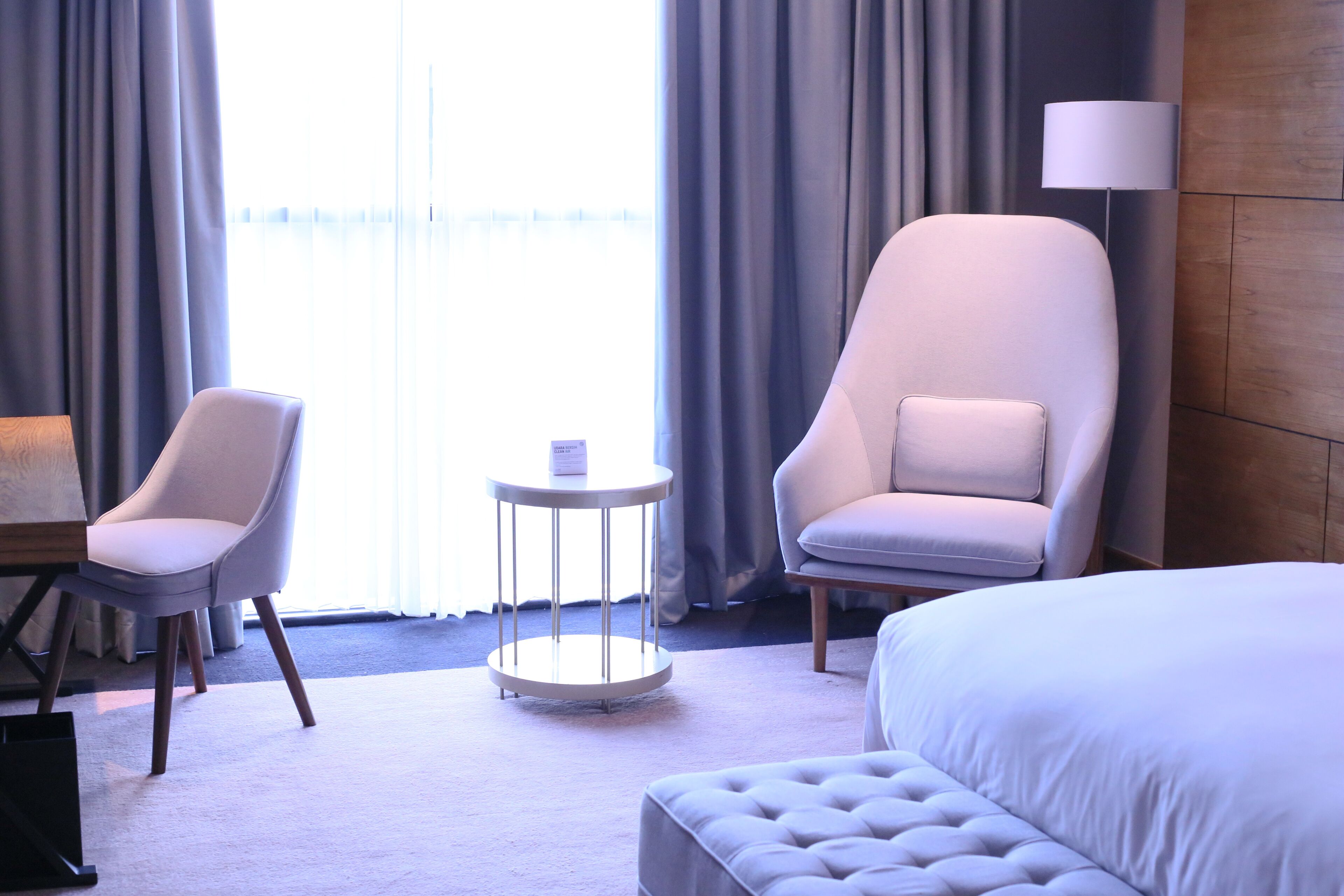 A hotel room with a bed, two armchairs, a small round table, a lamp, and a view of the outdoors through large windows.