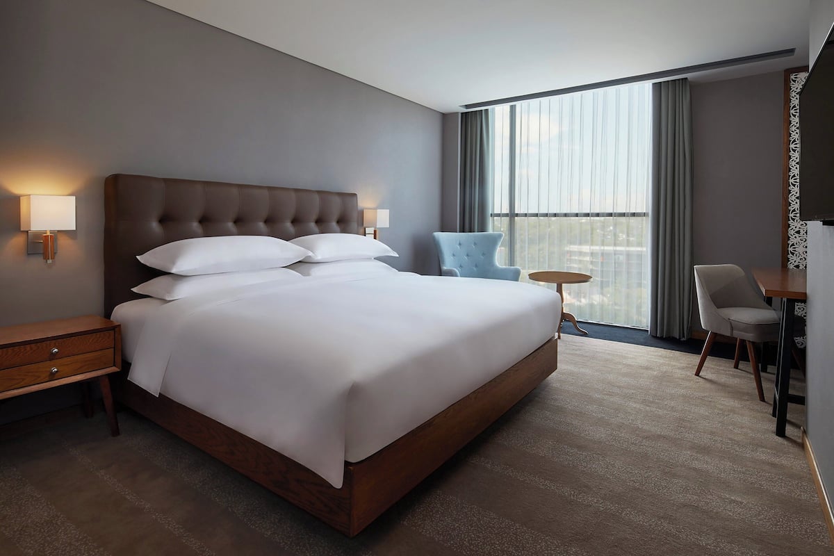 Une chambre d’hôtel moderne avec un grand lit, des tables de chevet, une chaise, une petite table ronde et une vue sur la ville grâce à de grandes fenêtres.