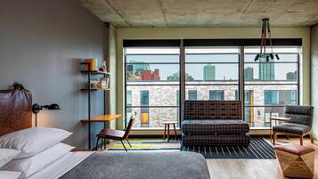 A modern hotel room with a large bed, a sofa, a chair, a small table, and a view of the city through large windows.