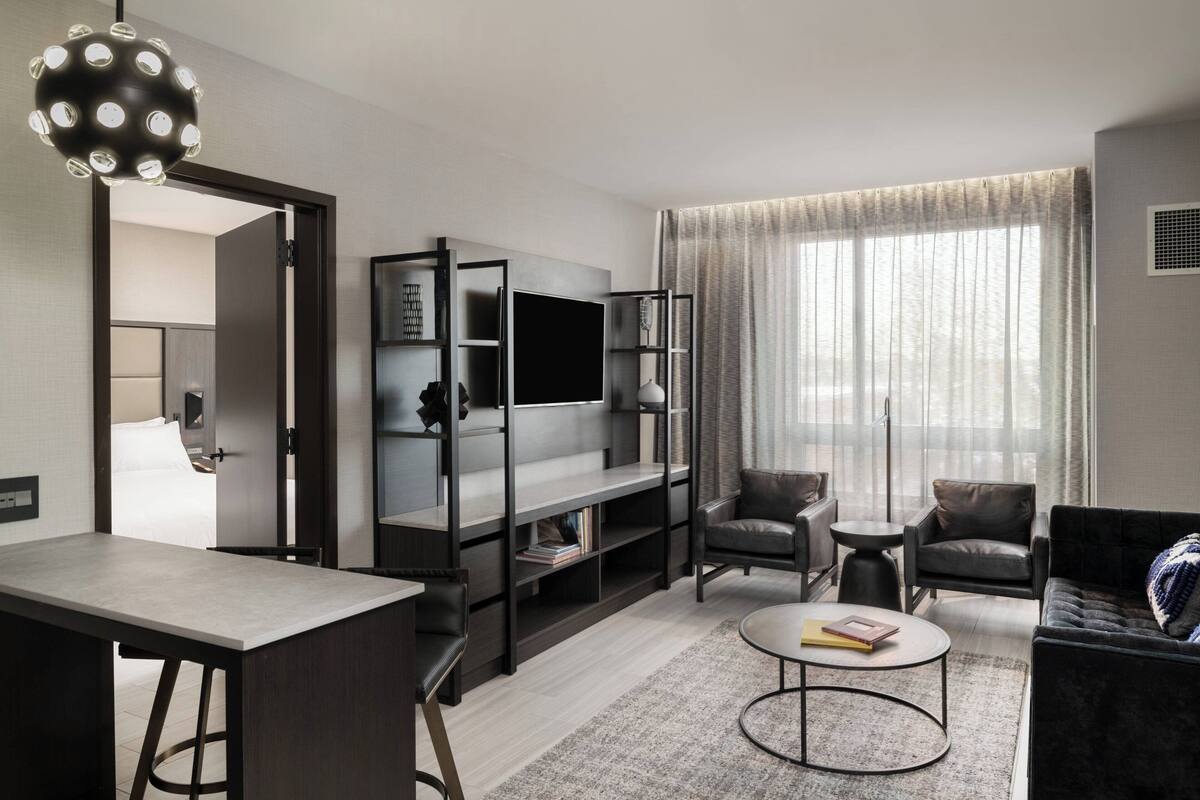 A modern living room with a flat-screen TV, dark leather sofas, a round coffee table, and a dining area with a bar stool.