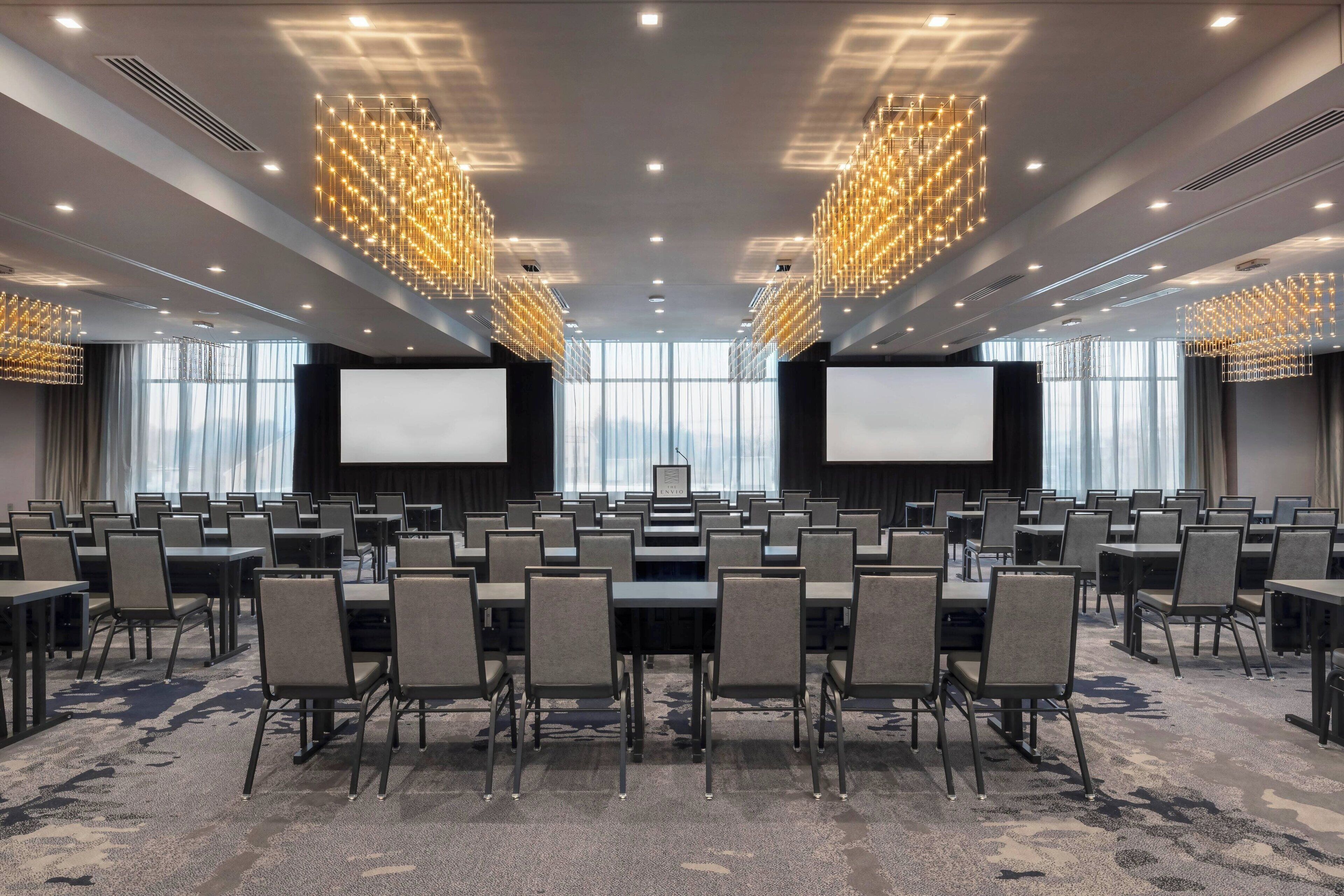 A conference room with rows of tables and chairs, large screens, and modern chandeliers.
