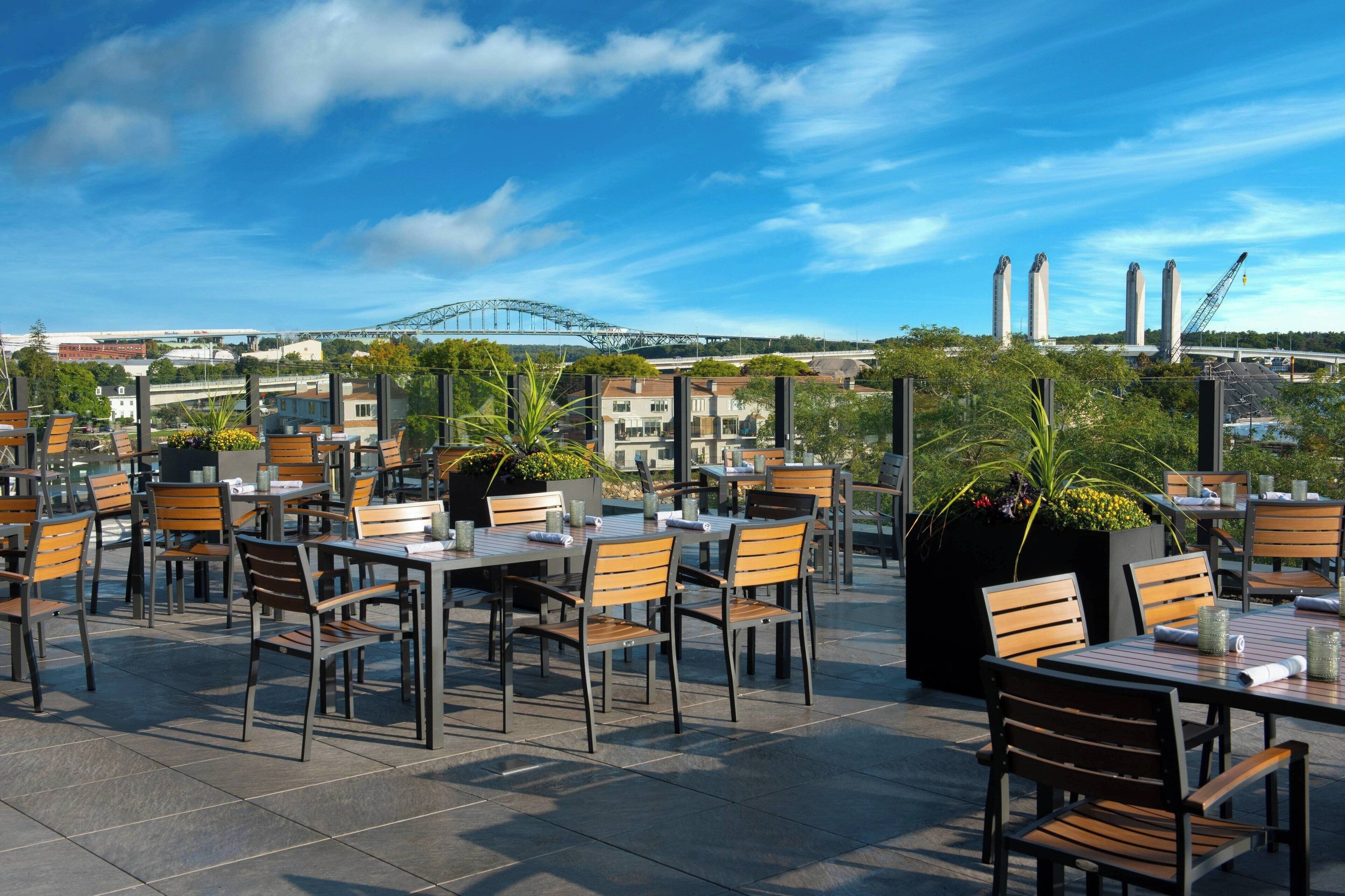 An outdoor dining area with tables and chairs, surrounded by potted plants and overlooking a cityscape with a bridge and industrial structures.