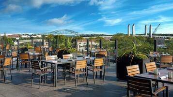 An outdoor dining area with tables and chairs, surrounded by potted plants and overlooking a cityscape with a bridge and industrial structures.