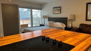 Une cuisine moderne avec un plan de travail en bois et une plaque de cuisson à induction, un lit avec une tête de lit et une grande fenêtre donnant sur une maison et des arbres.