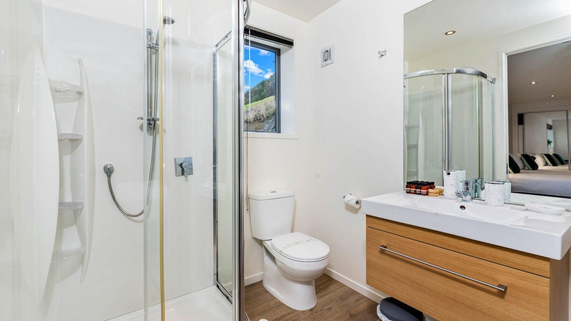 Un bagno moderno con cabina doccia in vetro, un water bianco, un mobile lavabo con specchio e vista su una camera da letto attraverso la finestra.