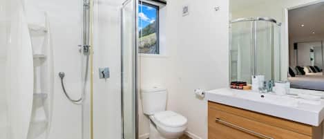 Un bagno moderno con cabina doccia in vetro, un water bianco, un mobile lavabo con specchio e vista su una camera da letto attraverso la finestra.