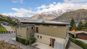 A modern two-story building with a flat roof, positioned on a hillside overlooking a lake and mountains. - Quaint Sanctuary on Quartz | Incredible Views | (Queenstown)