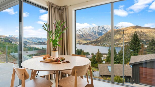 A dining area with a round table and chairs, a vase with a plant, and a view of mountains and a lake. - Quaint Sanctuary on Quartz | Incredible Views | (Queenstown)