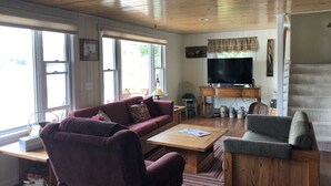 A cozy living room with a wooden ceiling, a large TV, a sofa, a coffee table, and a staircase. - Welcome to Heav'nsent (Swanville)