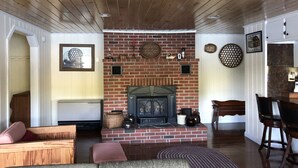 A cozy living room with a brick fireplace, wooden ceiling, and a dining area with a table and chairs. - Welcome to Heav'nsent (Swanville)