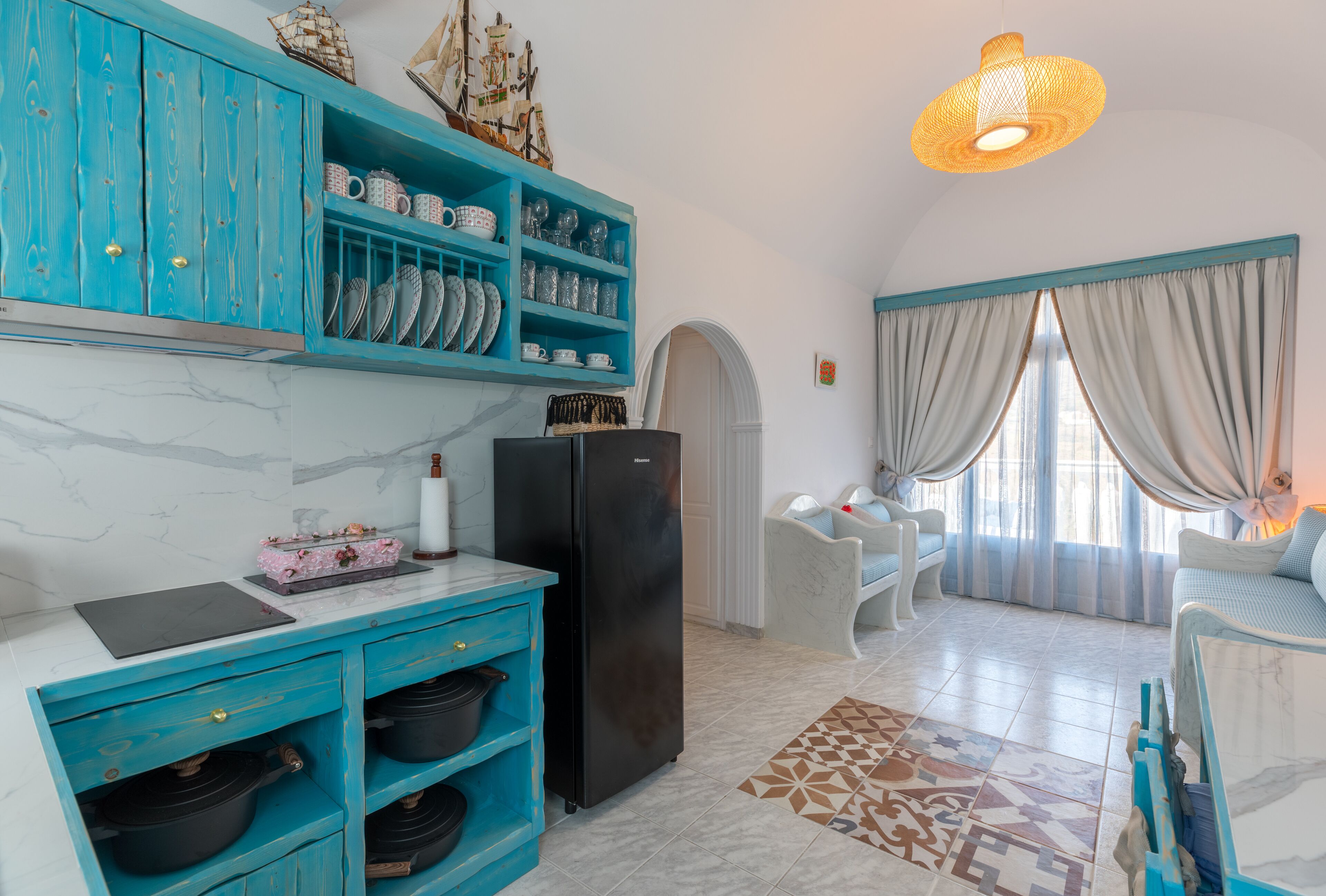 A compact kitchen with teal cabinetry, a black refrigerator, and a white countertop. There is a dining area with a white chair and a table, and a window with curtains.