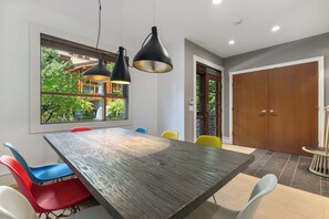 A dining area with a wooden table, colorful chairs, pendant lights, and a view of greenery outside.