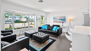A modern living room with a black leather sofa, matching ottoman, and a white coffee table. There is a large window with a view of the street and greenery outside.