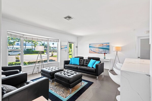 A modern living room with a black leather sofa, matching ottoman, and a white coffee table. There is a large window with a view of the street and greenery outside. - Entire Waterfront Home with Heated Pool, Dock & Sunset Views (Wilton Manors)
