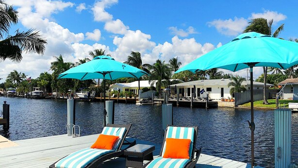 A waterfront deck with lounge chairs and umbrellas, a dock with boats, and a house in the background.