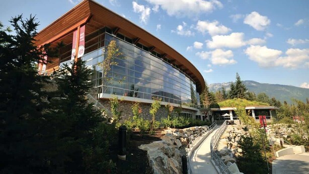 A modern building with a glass facade and a wooden roof, surrounded by greenery and mountains.