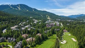 A golf course with a clubhouse, surrounded by residential buildings and dense forest, with mountains in the background.