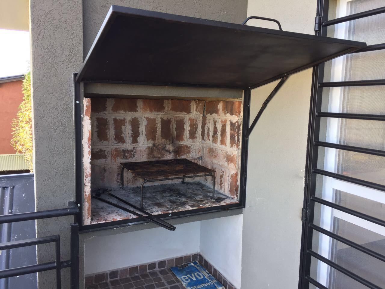Una parrilla pequeña al aire libre adosada a la pared exterior de un edificio, con una barandilla metálica y una ventana cerca.