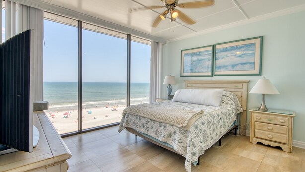 Een slaapkamer met een groot raam dat uitkijkt op een strand, een bed met een beddengoed met een patroon, een plafondventilator en een nachtkastje met een lamp.