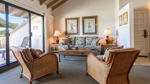 A cozy living room with wicker furniture, a coffee table, and two lamps. There are framed pictures on the wall and a sliding glass door leading outside. - Ocean-View Maui Kamaole Condo – A/C, Lanai, Walk to Beach & Pool! (Kihe)