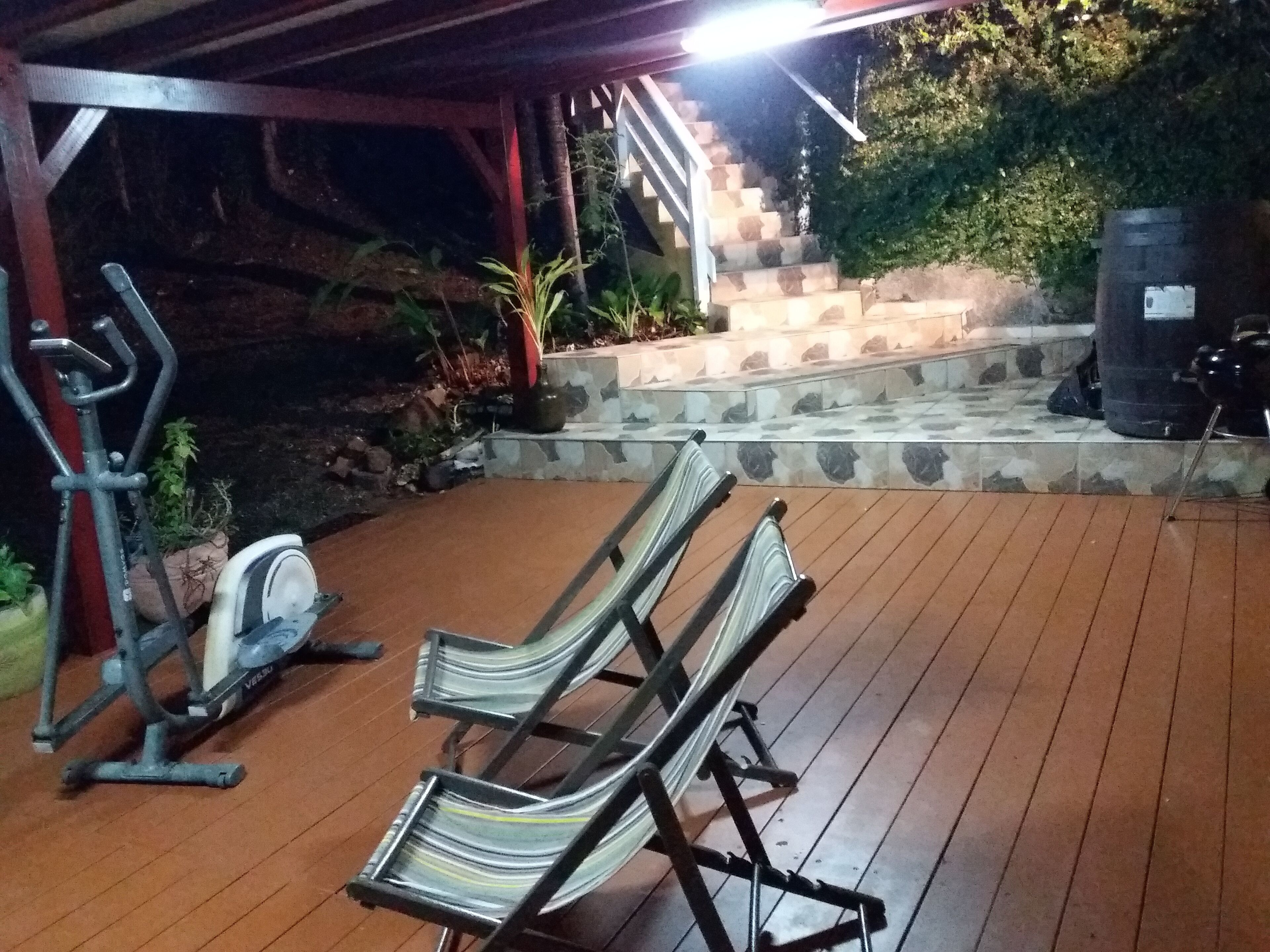 Un espacio al aire libre cubierto con terraza de madera, dos sillones, una bicicleta estática y una escalera que conduce a una plataforma de piedra con follaje.