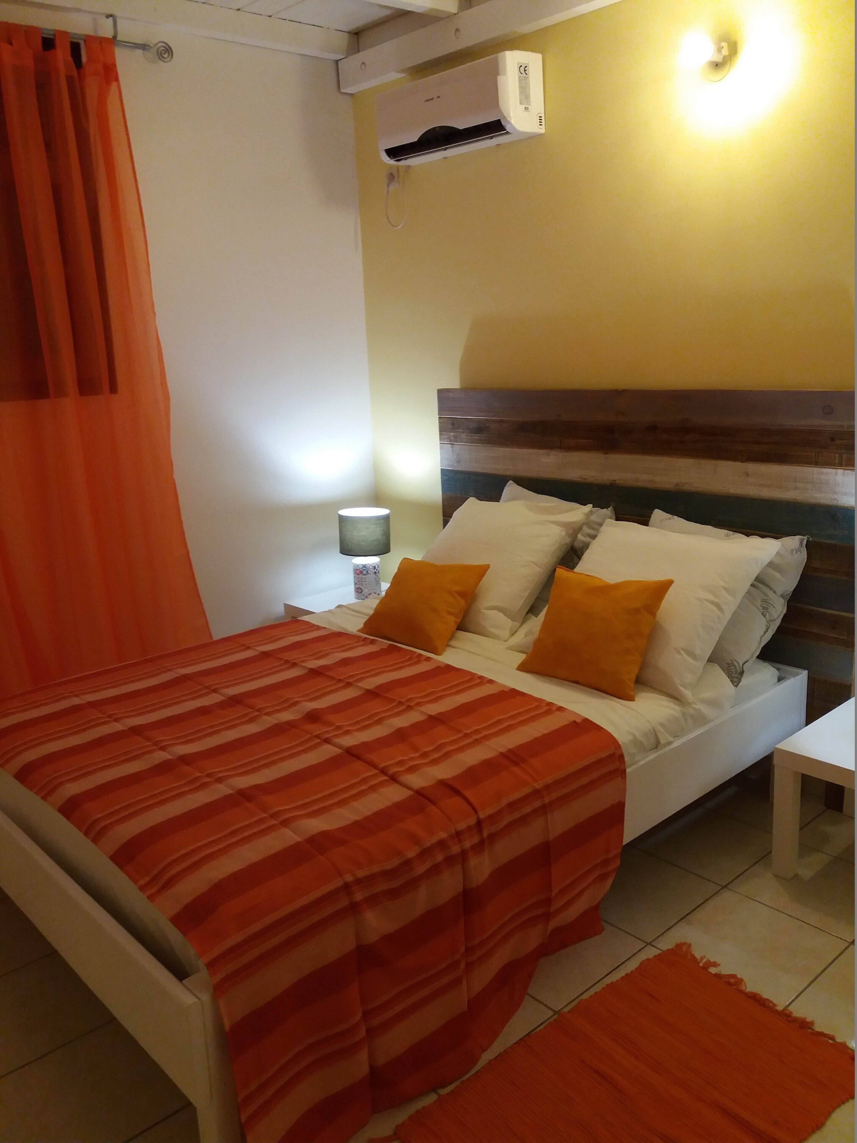 Un lit avec une couvre-lit orange à rayures, des oreillers blancs et un plaid orange. On y trouve une table de chevet avec une lampe, un climatiseur fixé au mur et un rideau.