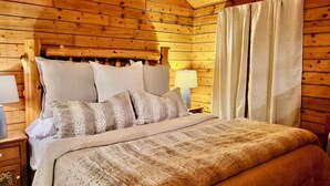 A wooden cabin-style bedroom with a bed, pillows, a nightstand, a lamp, and a window with curtains. - Tranquil Cabin- Perched on Mountain Top boasting jacuzzi for two and expansive deck with views! Trail & Cave on Property! (Eureka Springs)