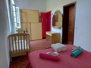 A room with a crib, a bed with rolled towels, a red wardrobe, a mirror, and a wooden door.