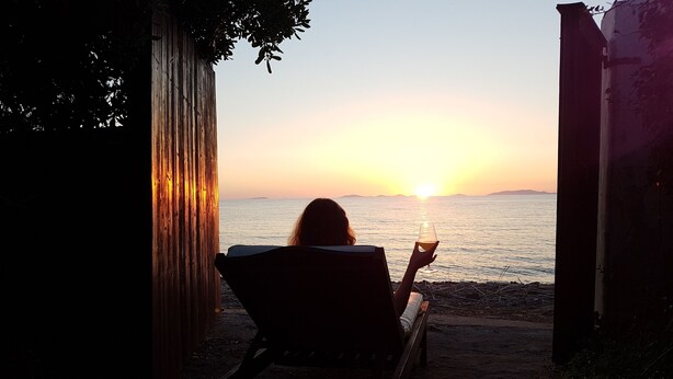 Une personne assise sur un banc, face à un coucher de soleil sur l’eau, tenant un verre à la main.