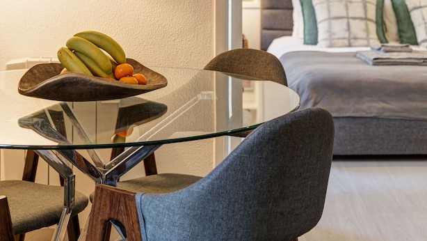 Un salon moderne avec une table à manger en verre, un canapé et un lit doté d’une tête de lit grise.