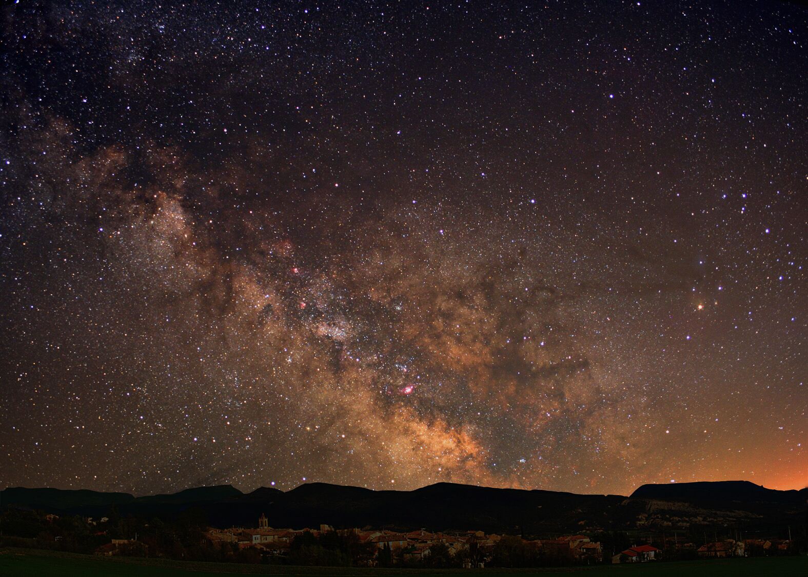 A night sky filled with stars and the Milky Way over a mountainous landscape with buildings at the base.