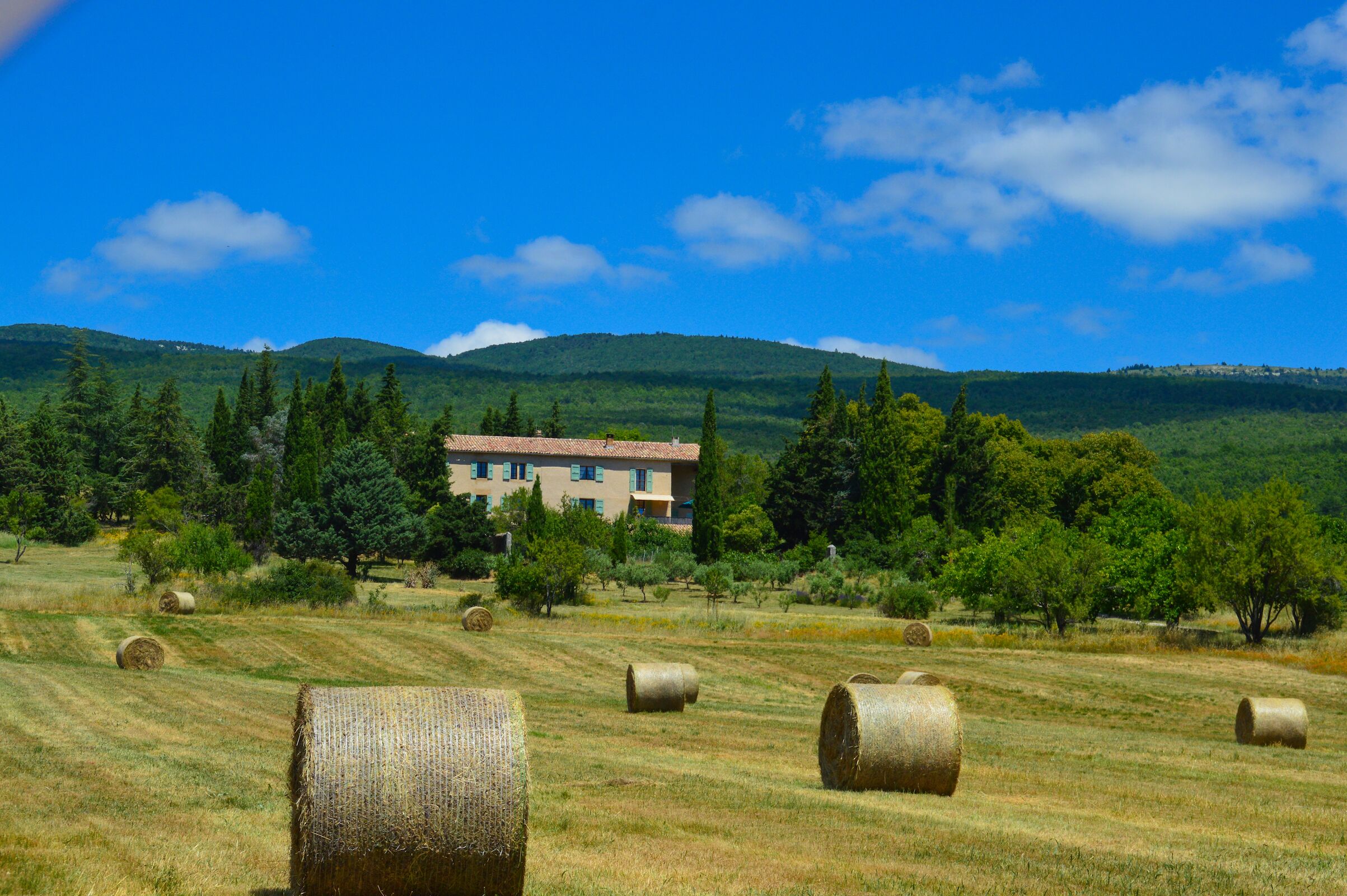 A rural landscape with hay bales, a building with a red roof, and green hills in the background.