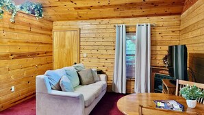 A cozy cabin-style room with a sofa, a round wooden table, and a television. - Tranquil Cabin- Perched on Mountain Top boasting jacuzzi for two and expansive deck with views! Trail & Cave on Property! (Eureka Springs)