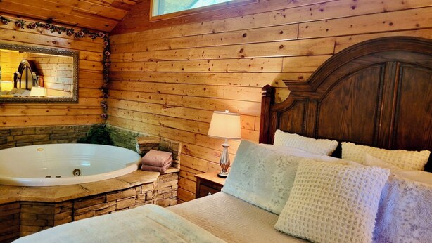 A wooden cabin bedroom with a large bed, a wooden headboard, a stone wall, a wooden ceiling, a window, and a bathroom with a bathtub and a mirror. - Tranquil Cabin- Perched on Mountain Top boasting jacuzzi for two and expansive deck with views! Trail & Cave on Property! (Eureka Springs)