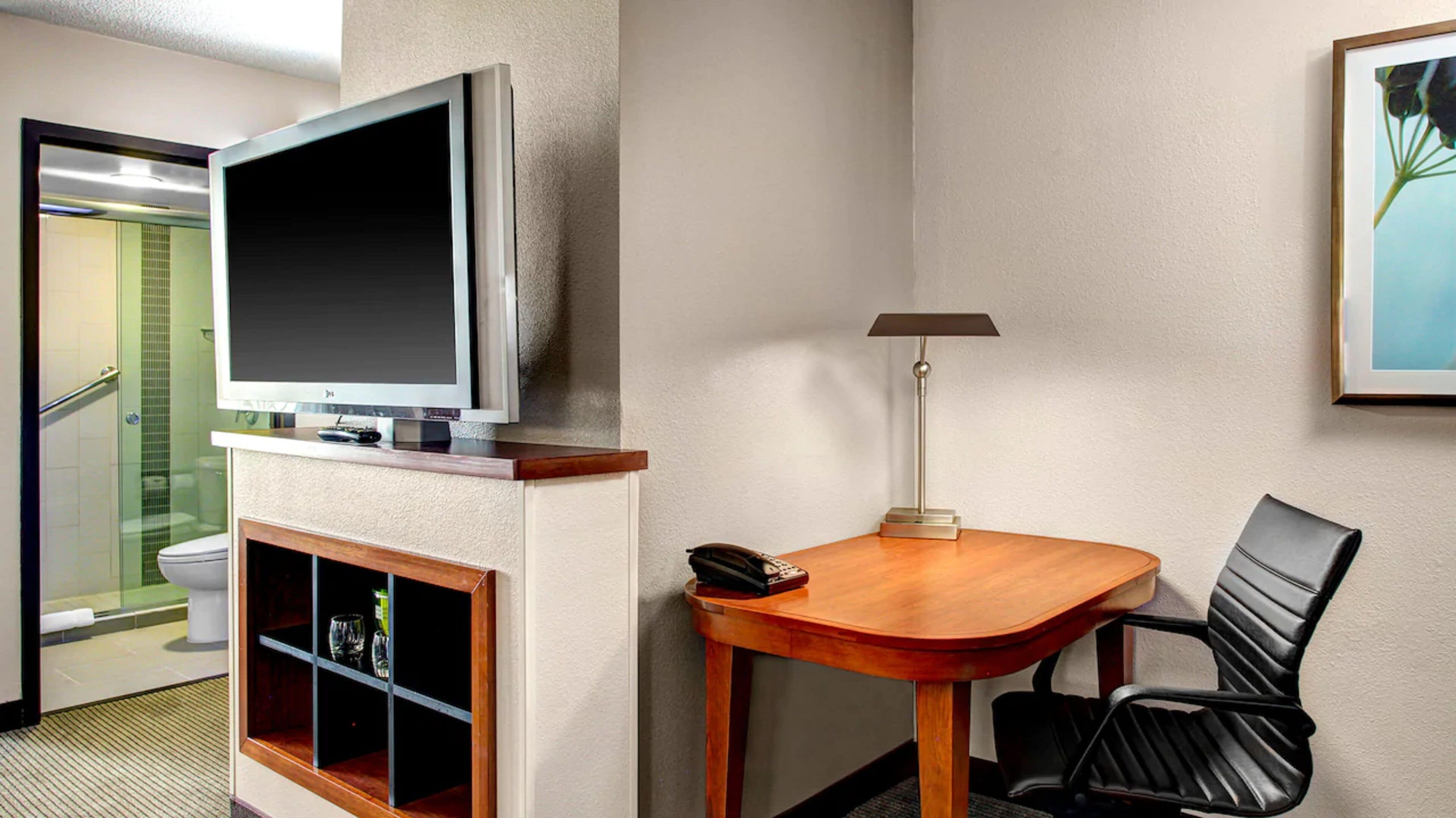 A hotel room with a television mounted on the wall, a wooden table with a lamp, a chair, and a shelf with glasses and a bottle.
