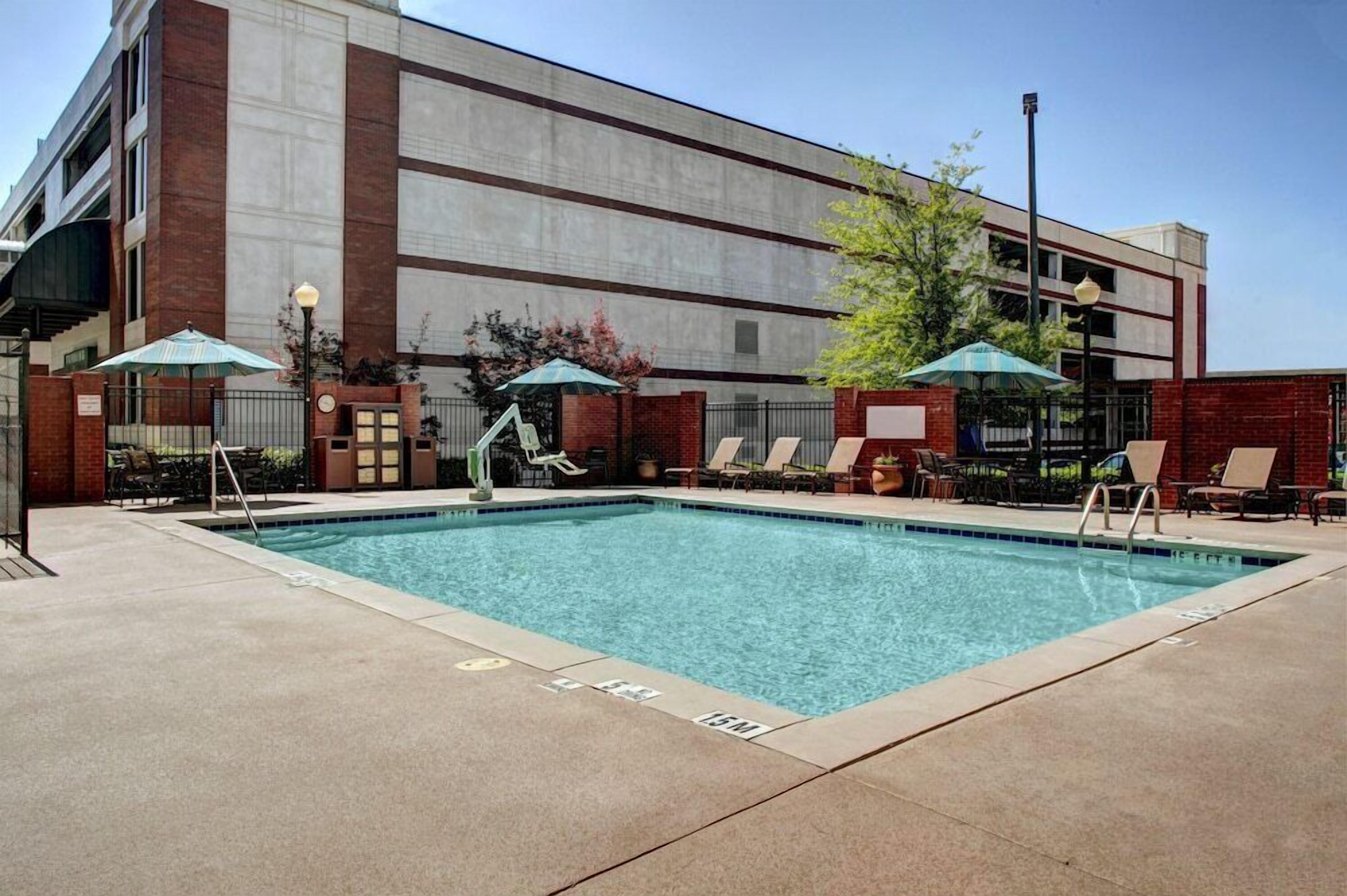 A rectangular pool with clear blue water, surrounded by lounge chairs and umbrellas, adjacent to a building with a brick facade and a flat roof.