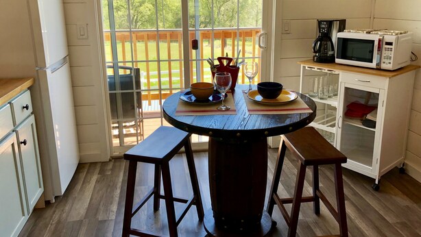 Une cuisine avec une table en bois, des tabourets, un réfrigérateur, un four à micro-ondes et une vue sur une terrasse bordée d’arbres.