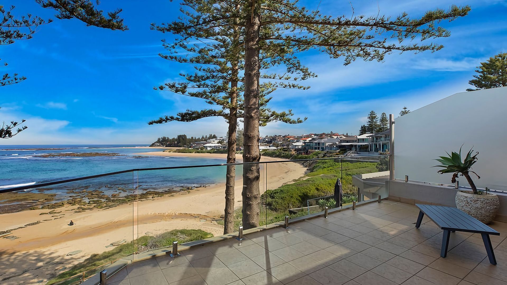 A balcony with a view of a beach and ocean, a potted plant, and a bench.