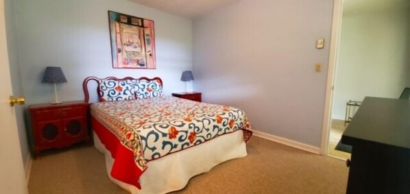 Un dormitorio con una cama, dos mesitas de noche, una lámpara, un cuadro en la pared y una puerta que da a otra habitación.