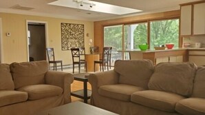 A living room with beige sofas, a dining area with chairs, a kitchen with cabinets, and a large window with a view of trees.