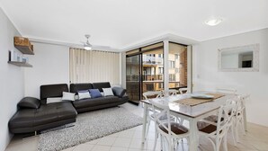 Een moderne woonkamer met een zwarte bank, een witte eettafel en stoelen, een plafondventilator en uitzicht op een balkon.