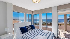 Un dormitorio con un ventanal grande que da a una playa, una cama con ropa de cama a rayas y una mesita de noche con una lámpara.